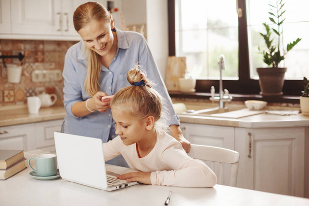 Mama hilft ihrer Tochter in der Küche mit einem Laptop im Internet bei den Hausaufgaben, spricht mit der Lehrerin. Fernunterricht, Online-Bildung, Heimschulkonzept - Foto, Bild