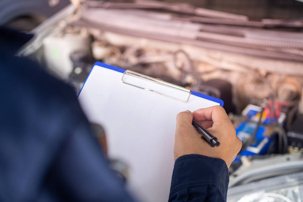 Nahaufnahmen des Werkstattmitarbeiters machen Notizen mit einem Klemmbrett, um den Motor des Autos zu überprüfen. - Foto, Bild
