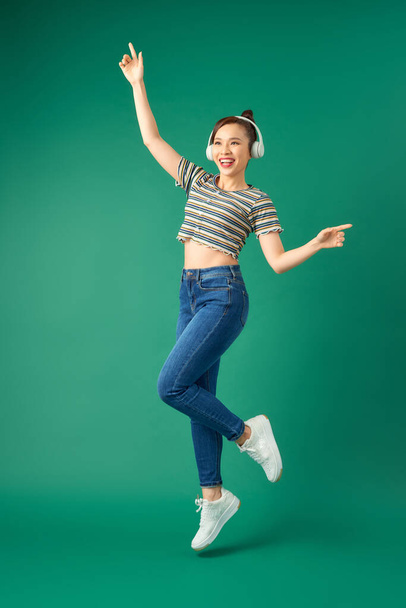 Full length portrait of young Asian woman in headphones listening to music dancing isolated green background - 写真・画像