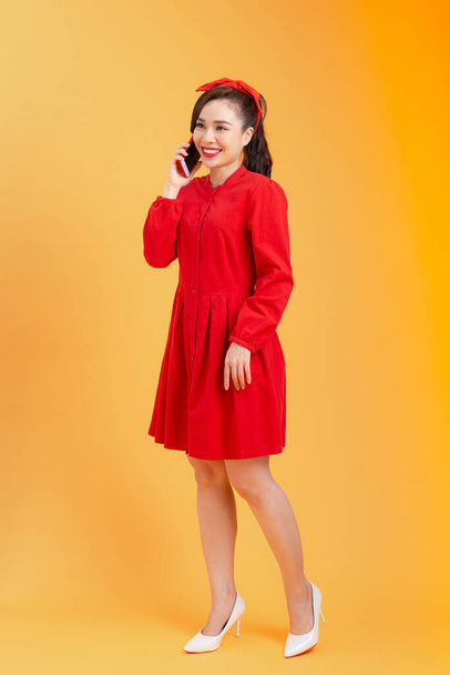 電話で話して赤いドレスで陽気な若いアジアの女性は、オレンジの背景の上に立っている。全長。全身. - 写真・画像