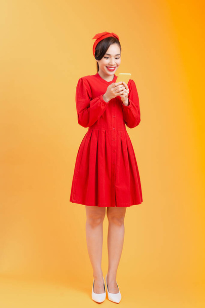 Πλήρες σώμα της ευτυχούς νεαρή Ασιάτισσα γυναίκα χρησιμοποιώντας το τηλέφωνο, φορώντας κόκκινο φόρεμα, ενώ στέκεται πάνω από πολύχρωμο φόντο. - Φωτογραφία, εικόνα