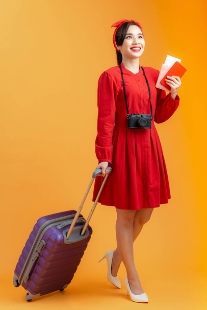 Cheerful young Asian woman holding suitcase, passport and flight ticket while standing isolated over orange background. - Photo, Image