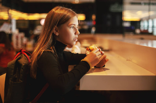 Güzel liseli kız bir kafede oturup hamburger yiyor. Lokantada abur cubur yiyen mutlu çocuk. Yan görünüm - Fotoğraf, Görsel