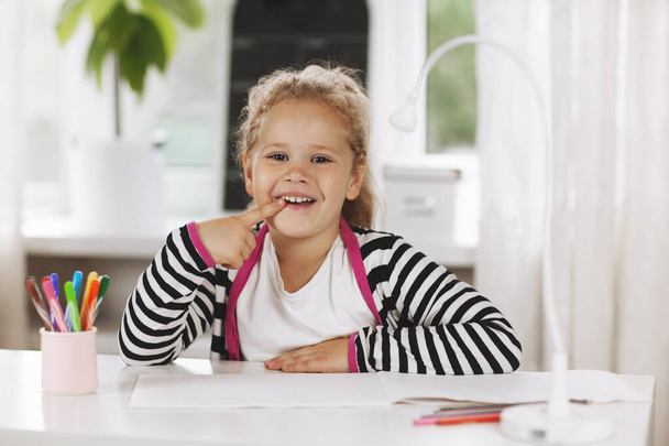 Wesoła dziewczynka siedząca przy stole i uśmiechnięta. Na stole jest szkicownik, kolorowe ołówki i lampa stołowa. Edukacja domowa, uczenie się przez Internet, odległość społeczna - Zdjęcie, obraz