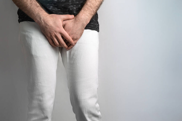 Valkoisiin farkkuihin pukeutunut mies, joka pitää sukupuolielimiä kädestä. Miesten sairaus. Käsite suojaa sukupuoliteitse tarttuvilta infektioilta. Kivessyöpä. - Valokuva, kuva