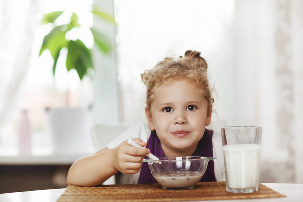 Чарівна дівчина їсть каші з молоком на сніданок і . На столі шоколадні пластівці, склянка молока. Здоровий сніданок, догляд за дітьми. Простір для тексту
 - Фото, зображення