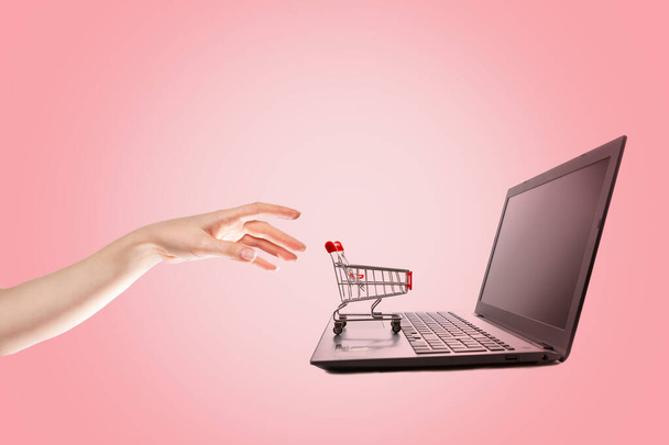 Shopping. A woman's hand reaches for a mini grocery cart on her laptop. Pink background. Copy space. The concept of online shopping. - Photo, Image