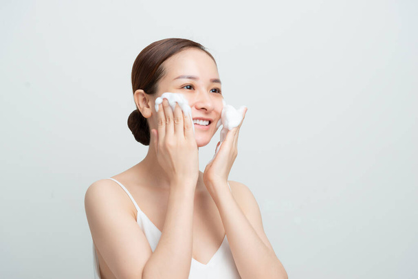 Sonriendo hermosa chica asiática usando limpiador de espuma. Concepto de lavado facial aislado sobre fondo blanco. - Foto, Imagen