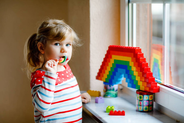 Cute little toddler girl by window create rainbow with colorful plastic blocks during pandemic coronavirus quarantine. Children made and paint rainbows around the world as sign. Kid eating lollipop. - Photo, Image