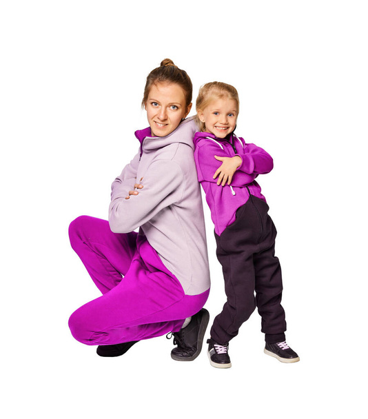 Glimlachend blond meisje en vrouw in stijlvolle jumpsuits met capuchon poserend over grijze achtergrond. Leuke dochter van 4-5 jaar oud die haar moeder knuffelt. Mock up kopieerruimte lifestyle concept. - Foto, afbeelding
