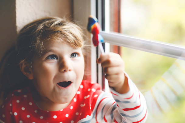 Мила маленька дівчинка, яка сидить біля вікна в дощовий день під час пандемії коронавірусу карантину. Чарівна дитина їсть і облизує веселку кольоровим льодяником
 - Фото, зображення