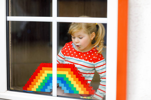 Cute little toddler girl by window create rainbow with colorful plastic blocks during pandemic coronavirus quarantine. Children made and paint rainbows around the world as sign. - Photo, Image
