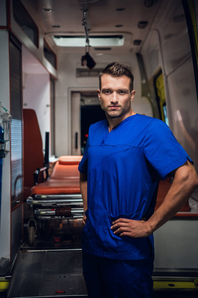 Handsome man in a medical uniform looking at the camera, ambulance car in the background - Fotó, kép