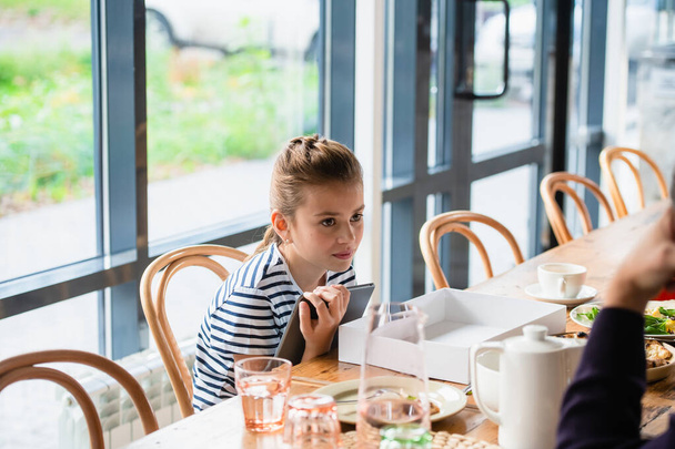 Ein hübsches Mädchen sitzt an einem Tisch mit einem Tablet. Verschwommenes Rückgrat - Foto, Bild