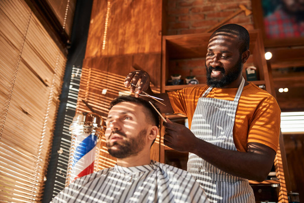Улыбающийся парикмахер стрижет волосы клиента ножницами в парикмахерской - Фото, изображение