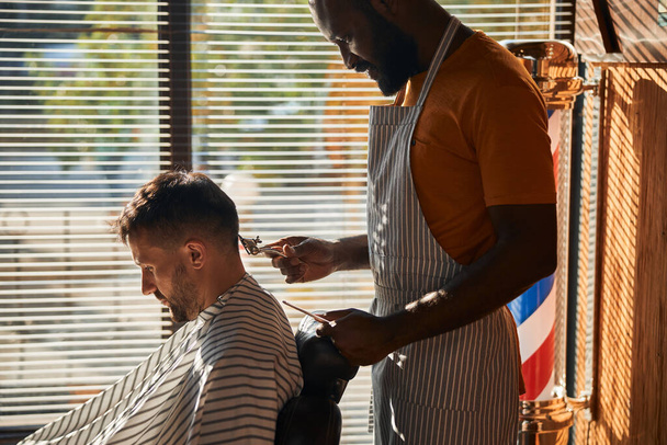 Όμορφος αρσενικός κουρέας που κόβει τα μαλλιά του πελάτη με μεταλλικό ψαλίδι μαλλιών - Φωτογραφία, εικόνα