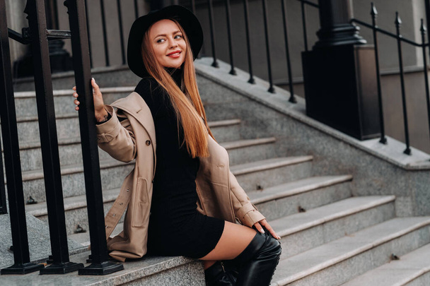 Stylish young woman in a beige coat in a black hat on a city street. Women's street fashion. Autumn clothing.Urban style. - Photo, Image