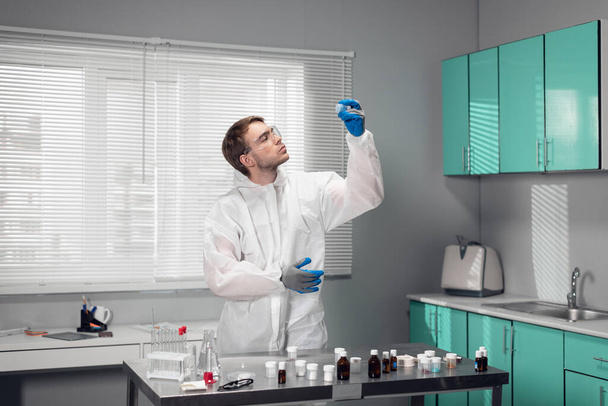 Un científico en gafas protectoras y monos blancos de pie en la mesa en el laboratorio de ciencias, estudiando nuevas sustancias - Foto, Imagen