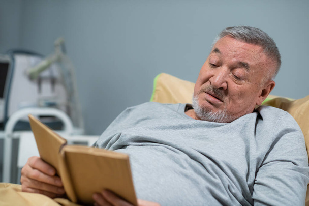 A male patient in his mid sixties reading a book in a hospital ward - Zdjęcie, obraz
