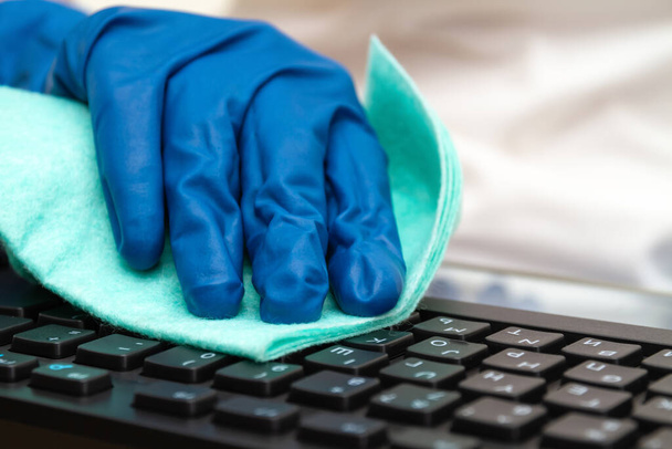 Girl's hand in blue protective glove cleans keyboard with a cloth for prevention coronavirus, bacteria, viruses and germs. - 写真・画像