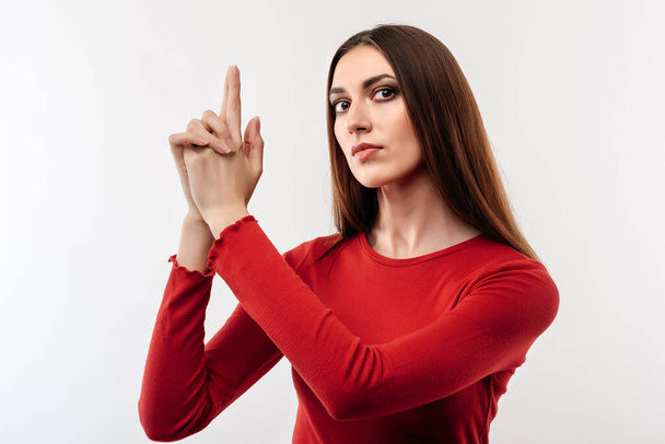 Retrato de una joven morena confiada en suéter rojo casual tomándose de las manos como un arma. Gente emociones sinceras concepto de estilo de vida. Captura de estudio, fondo blanco - Foto, Imagen