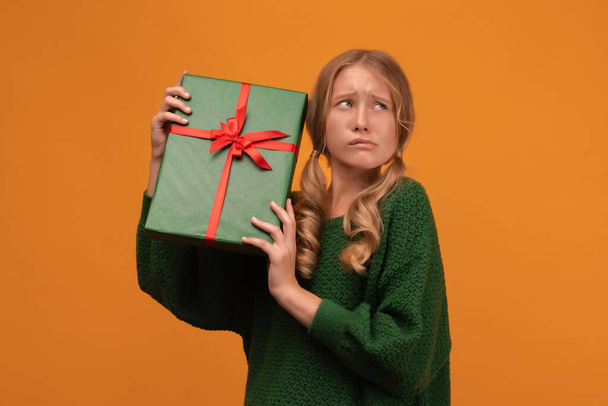 Wizerunek czarującej blondynki 12-14 lat w ciepłym zielonym swetrze, trzymającej pudełko z czerwoną kokardą. Zdjęcia studyjne, żółte tło, odizolowane. Nowy Rok Dzień Kobiet urodziny koncepcja wakacje - Zdjęcie, obraz
