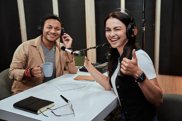 Portait των δύο χαρούμενων ραδιοφωνικών παρουσιαστών, άνδρας και γυναίκα χαμογελώντας στην κάμερα, πίνοντας καφέ ή τσάι, ενώ ετοιμάζεται για μια ζωντανή παράσταση στο στούντιο. Εστίαση στη γυναίκα παρουσιάστρια που δείχνει τους αντίχειρες - Φωτογραφία, εικόνα