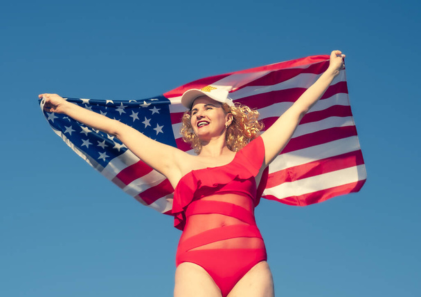 Mulher loira alegre bonita segurando uma bandeira americana na praia. Uma mulher de meia-idade de biquíni vermelho é patrioticamente entusiasta. Viajar. Bandeira dos EUA - Foto, Imagem