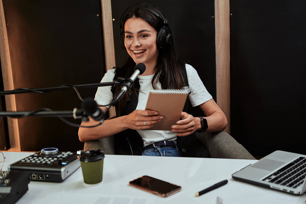 Portait of attractive young female radio host holding a script, looking emotional while speaking in microphone, moderating a live show - Фото, изображение