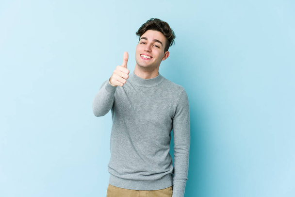 Νεαρός καυκάσιος άνδρας απομονωμένος σε μπλε φόντο χαμογελώντας και υψώνοντας τον αντίχειρα - Φωτογραφία, εικόνα