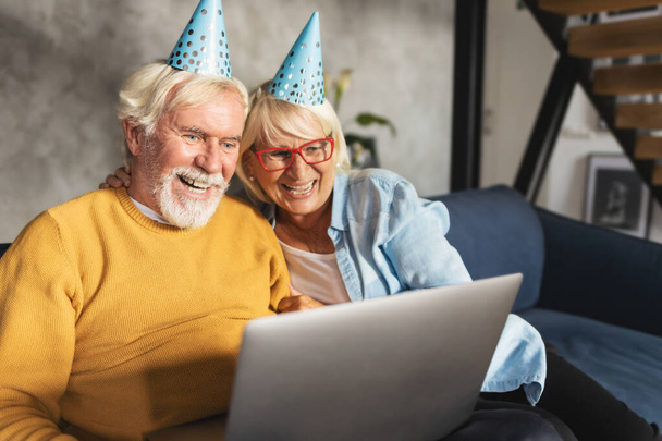 Веселая современная кавказская пожилая пара, сидя дома на диване, делает видеозвонок на ноутбуке на день рождения или юбилей. Концепция поздравления с праздником видеоконференции - Фото, изображение