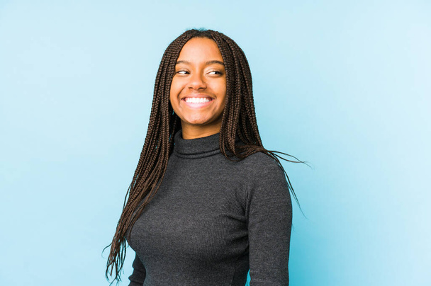 Νεαρή Αφροαμερικανή γυναίκα απομονωμένη σε μπλε φόντο κοιτάζει πέρα χαμογελαστή, χαρούμενη και ευχάριστη. - Φωτογραφία, εικόνα
