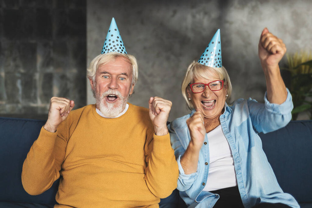 Portrait of funny senior couple in birthday caps having video call for celebration. Videoconference holiday greetings concept - Foto, Imagem