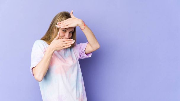 Young blonde woman isolated on purple background blink at the camera through fingers, embarrassed covering face. - Photo, Image