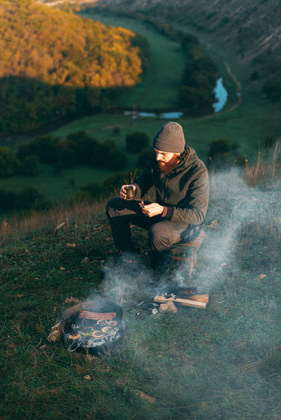 Jeune homme barbu sur une colline est assis près d'un gril avec un café - Photo, image