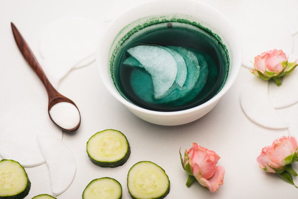 bowl with cleansing lotion and cotton pads near sliced cucumber, tea roses, and wooden spoon with cosmetic cream on white - Photo, Image