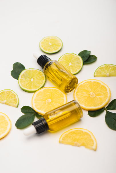 jugosas rodajas de limón y lima cerca de botellas con aceite esencial de cítricos y hojas de rosa sobre blanco - Foto, imagen