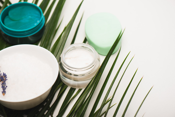 containers with cosmetic cream and scrub, soap and bowl with lavender twig near palm leaves on white - Foto, afbeelding