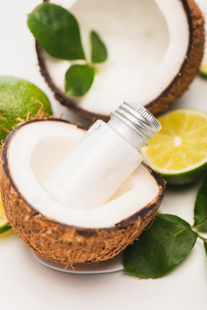 bottle of lotion in coconut half near lime, and rose leaves on blurred white background - Photo, Image