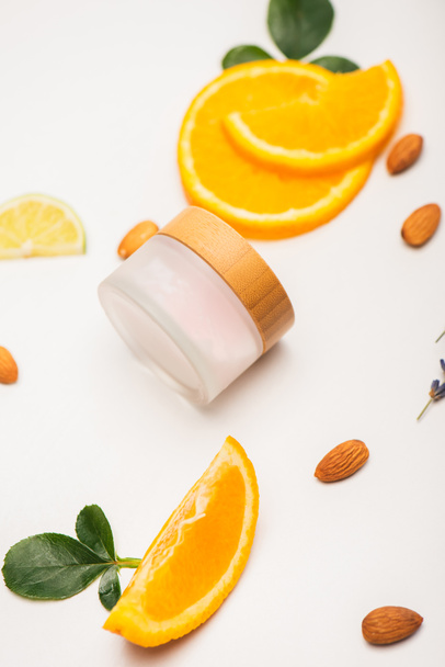 container met zelfgemaakte cosmetische crème bij verse sinaasappelschijfjes, amandelen en rozenblaadjes op wit - Foto, afbeelding