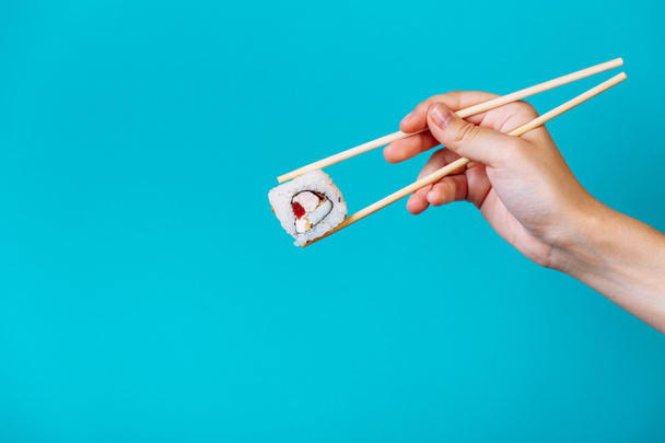 女性の手には青い背景に木の箸でおいしい寿司ロール巻きが閉じられます。キャプションとテキストの場所. - 写真・画像