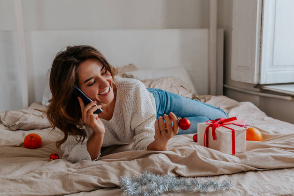 молодая девушка лежит на кровати и говорит по телефону. Рождественское настроение - Фото, изображение