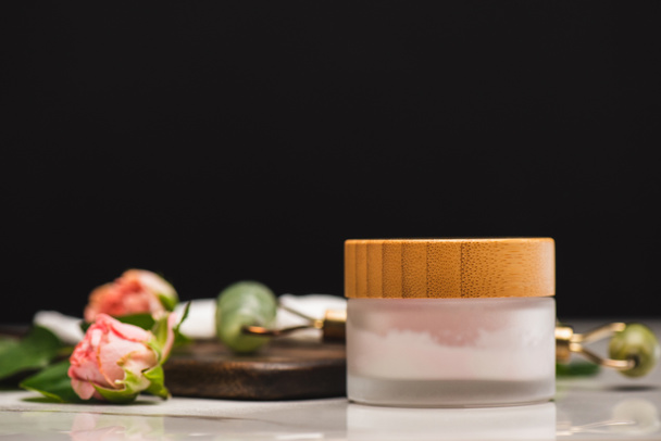 container with homemade cosmetic cream near jade roller and tea roses on chopping board on blurred background isolated on black - Foto, afbeelding