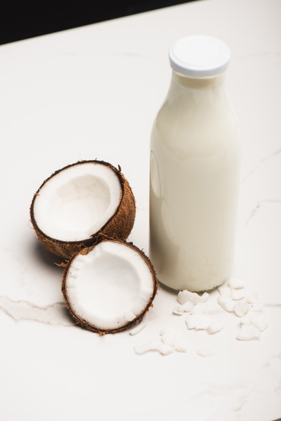 fles kokosmelk bij helften en vlokken op wit oppervlak geïsoleerd op zwart - Foto, afbeelding