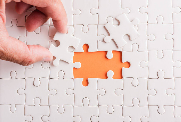 Top view flat lay of hand-holding last piece white paper jigsaw puzzle game last pieces put to place for solve problem complete mission, studio shot on an orange background, quiz calculation concept - Photo, Image