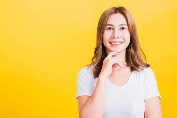 Portrait Asian Thai beautiful young woman wearing white t-shirt standing chin handle relaxed thinking about something about the question studio shot, isolated on yellow background with copy space - Foto, imagen