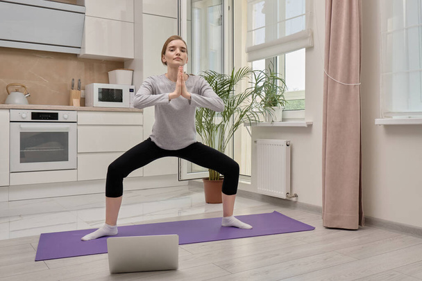 A blonde woman does yoga at home on a yoga Mat and looks at the camera. - Фото, изображение