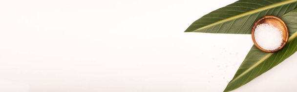верхний вид морской соли в деревянной чаше и зеленые листья на белом фоне с копировальным пространством, баннер - Фото, изображение