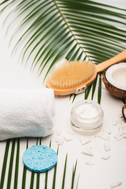 Кокосовая половина, косметический крем, полотенце, массажная щетка и губка возле пальмовых листьев на белом - Фото, изображение