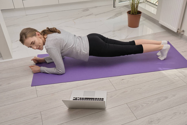A young blonde woman in a plank pose, athletic body, looks at the laptop screen. - Photo, Image